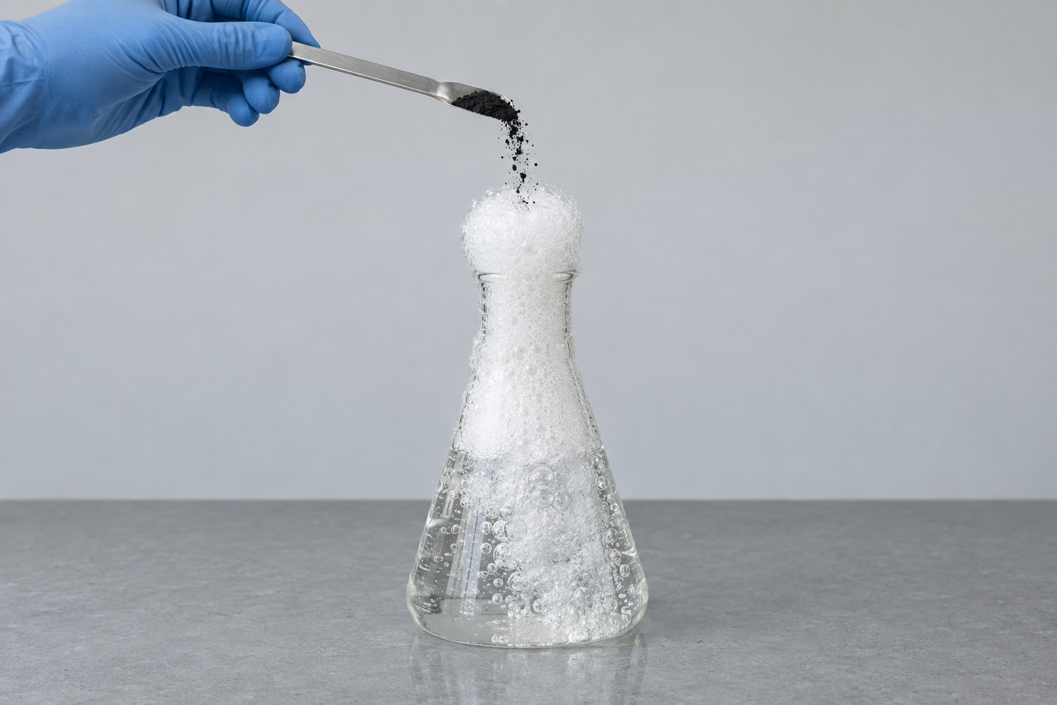 A gloved hand tips fine black powder from a spatula into an Erlenmeyer flask of colorless liquid. A tall column of dense white froth erupts up out of the flask's neck.