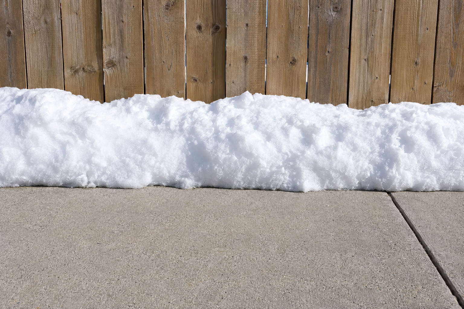 A snowbank in full sunshine beside a cleared path, with dry pavement and no meltwater anywhere.