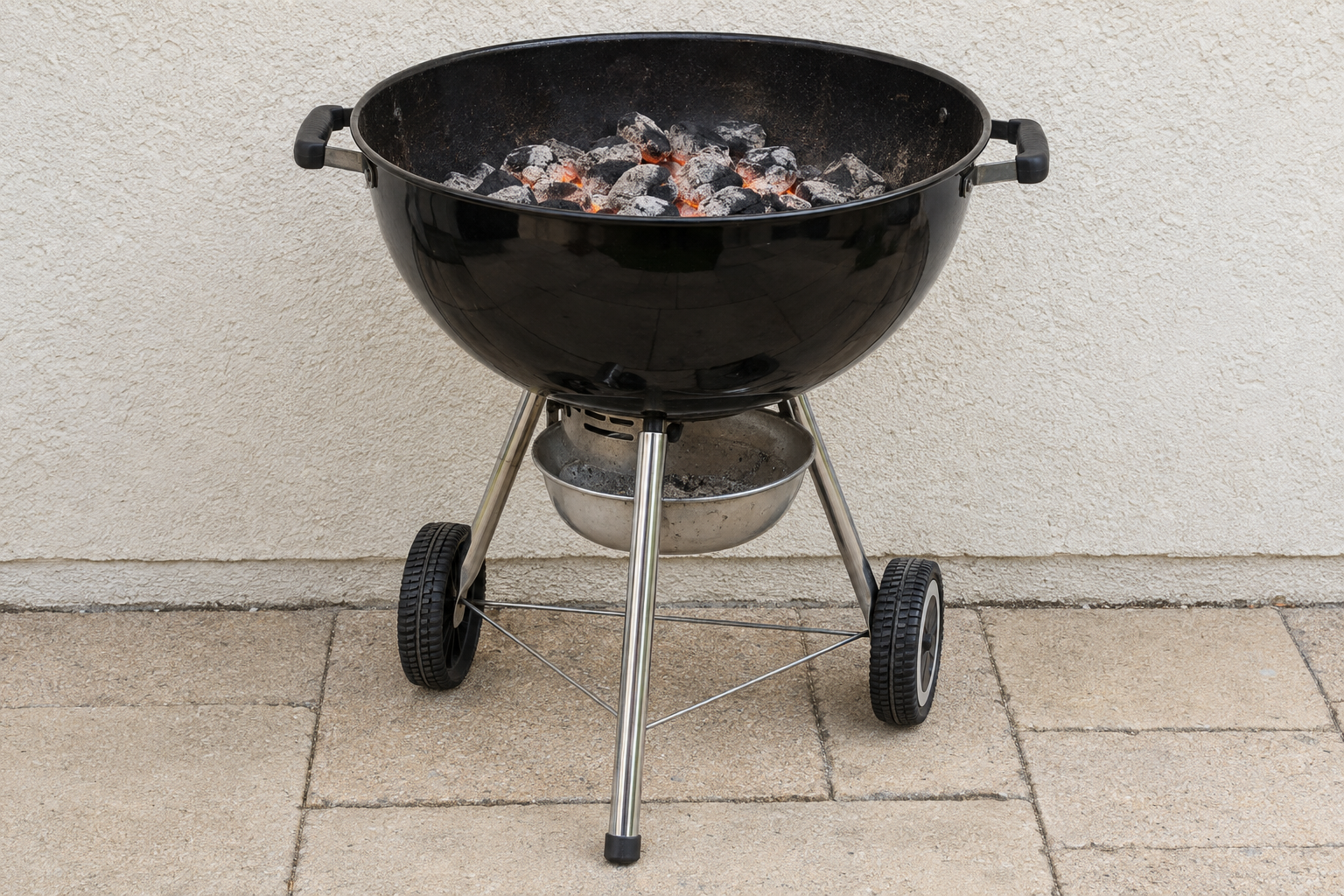 A kettle charcoal grill with a bed of coals, standing on flat paving.
