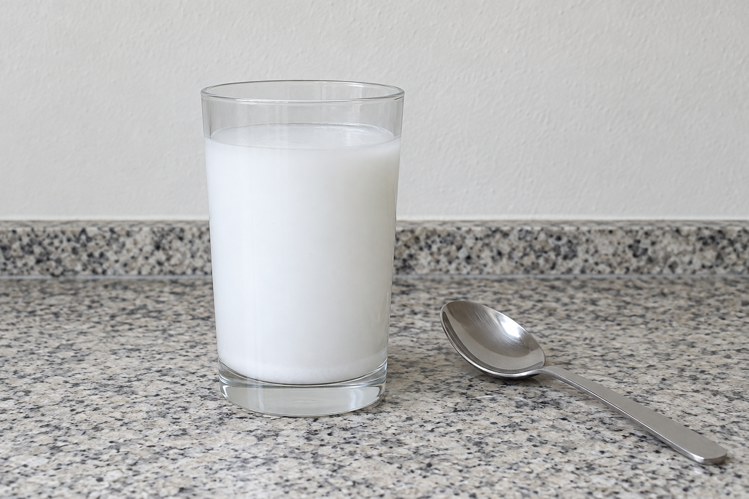 A clear drinking glass on a plain kitchen counter holding an opaque white mixture of fine solid stirred through water, with a thin layer of the white solid settled on the bottom of the glass. A metal teaspoon lies on the counter beside the glass.